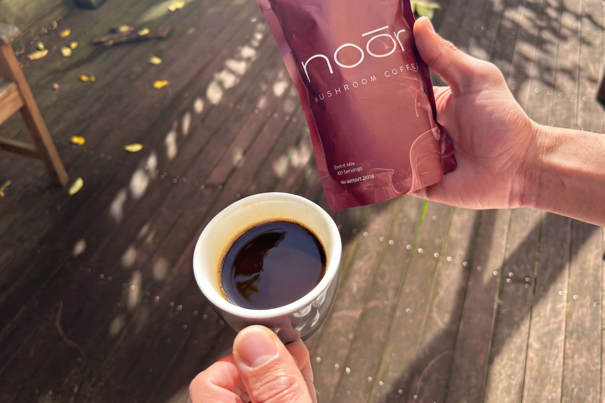 A person holds a white cup of black coffee and a pouch of Noor Mushroom Coffee outdoors on a wooden deck.