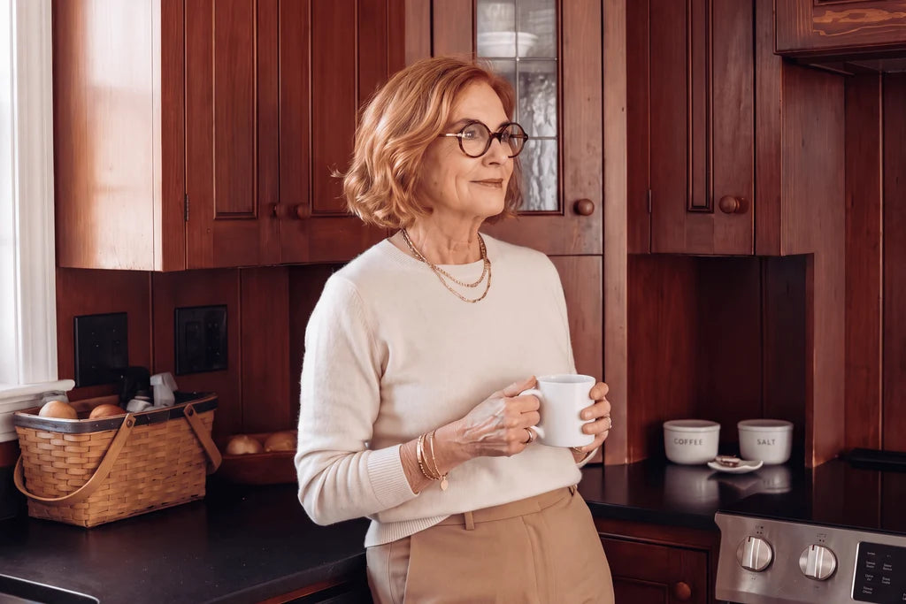 An older woman with short blonde hair and glasses stands in a warm wood kitchen, holding a white mug. She is wearing a cream knit sweater and tan pants.