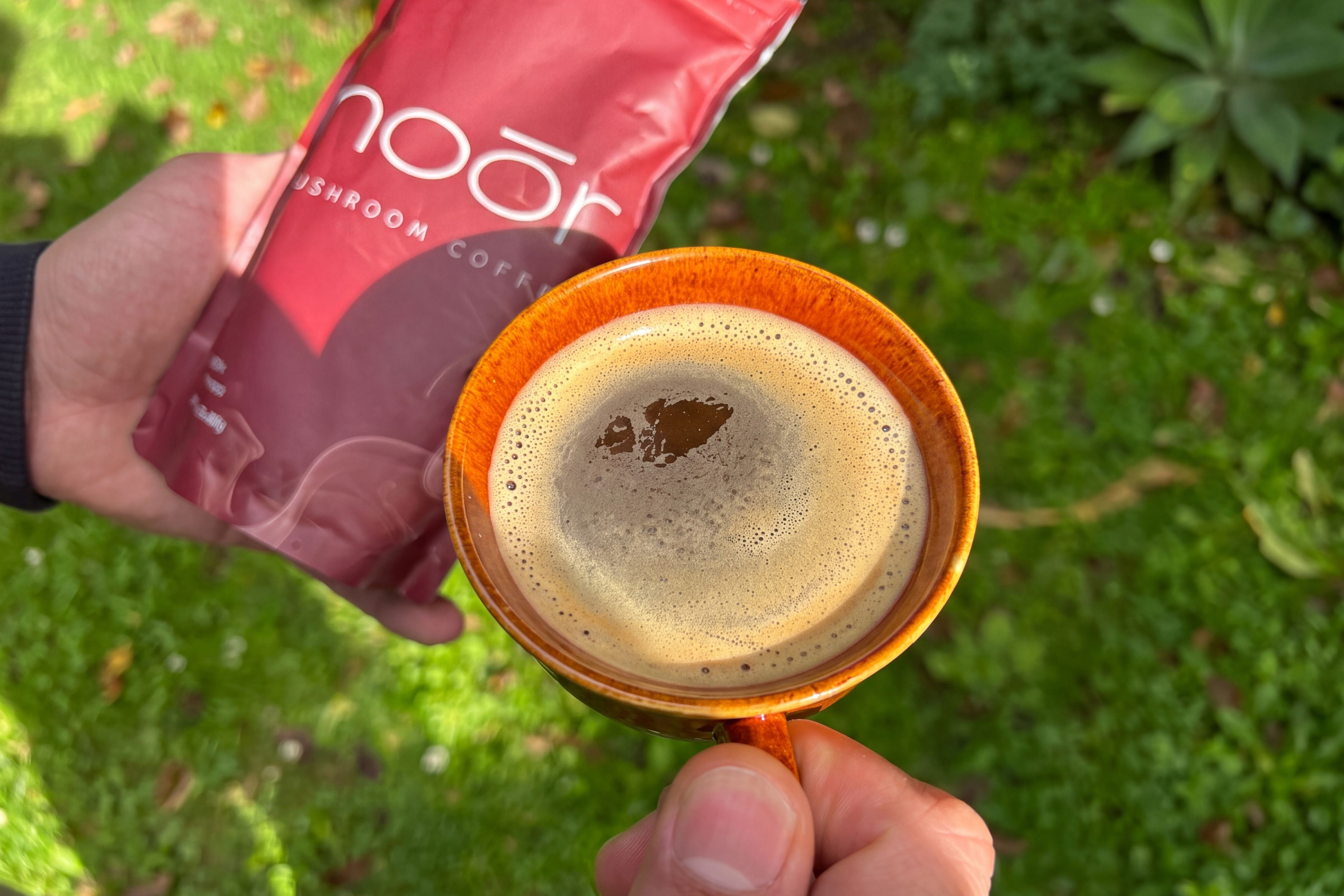A person holds a bright orange ceramic cup filled with espresso and foam in an outdoor garden setting, while holding a red pouch of Noór mushroom coffee in the other hand.