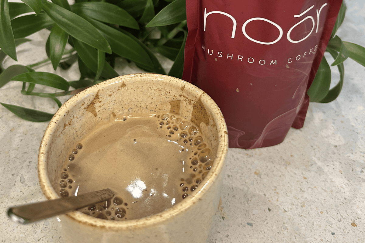 A ceramic mug filled with brewed mushroom coffee and a spoon, with a red pouch labeled 'noor mushroom coffee' standing behind it on a light speckled countertop, with green leaves in the background.
