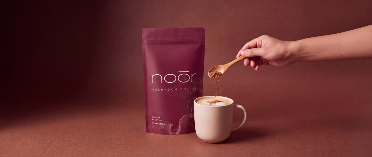 A hand holding a wooden spoon sprinkles powder over a white cup of coffee next to a dark magenta stand-up pouch labeled Noōr on a warm brown background.